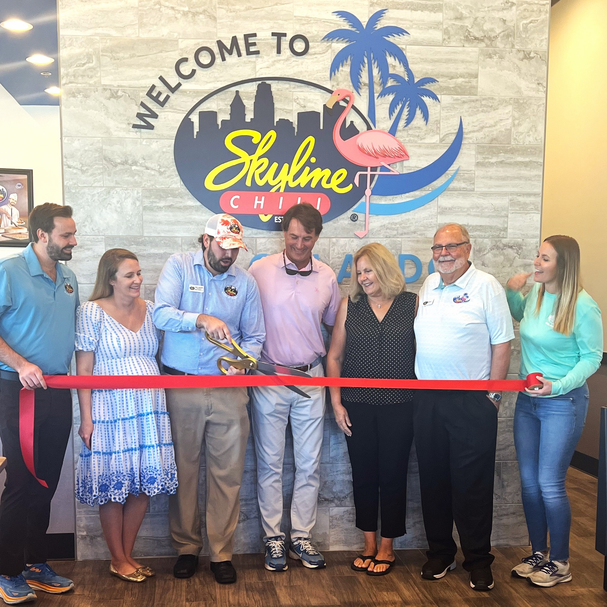 Image of 7 individuals standing in front of a Welcome to Skyline Chili Orlando sign, cutting a red ribbon for opening day